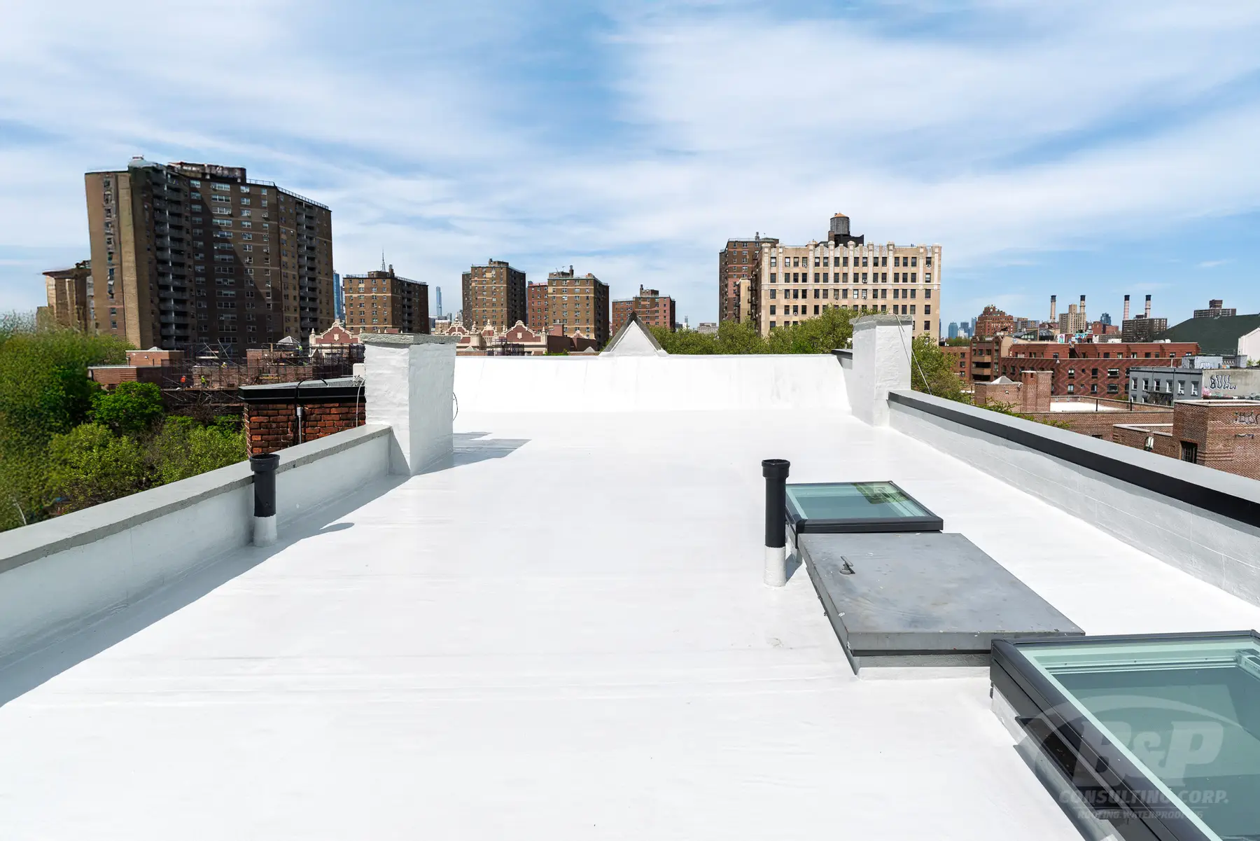 A finished white flat roof with new skylights on a Brooklyn rowhouse, apartment towers and blue sky behind